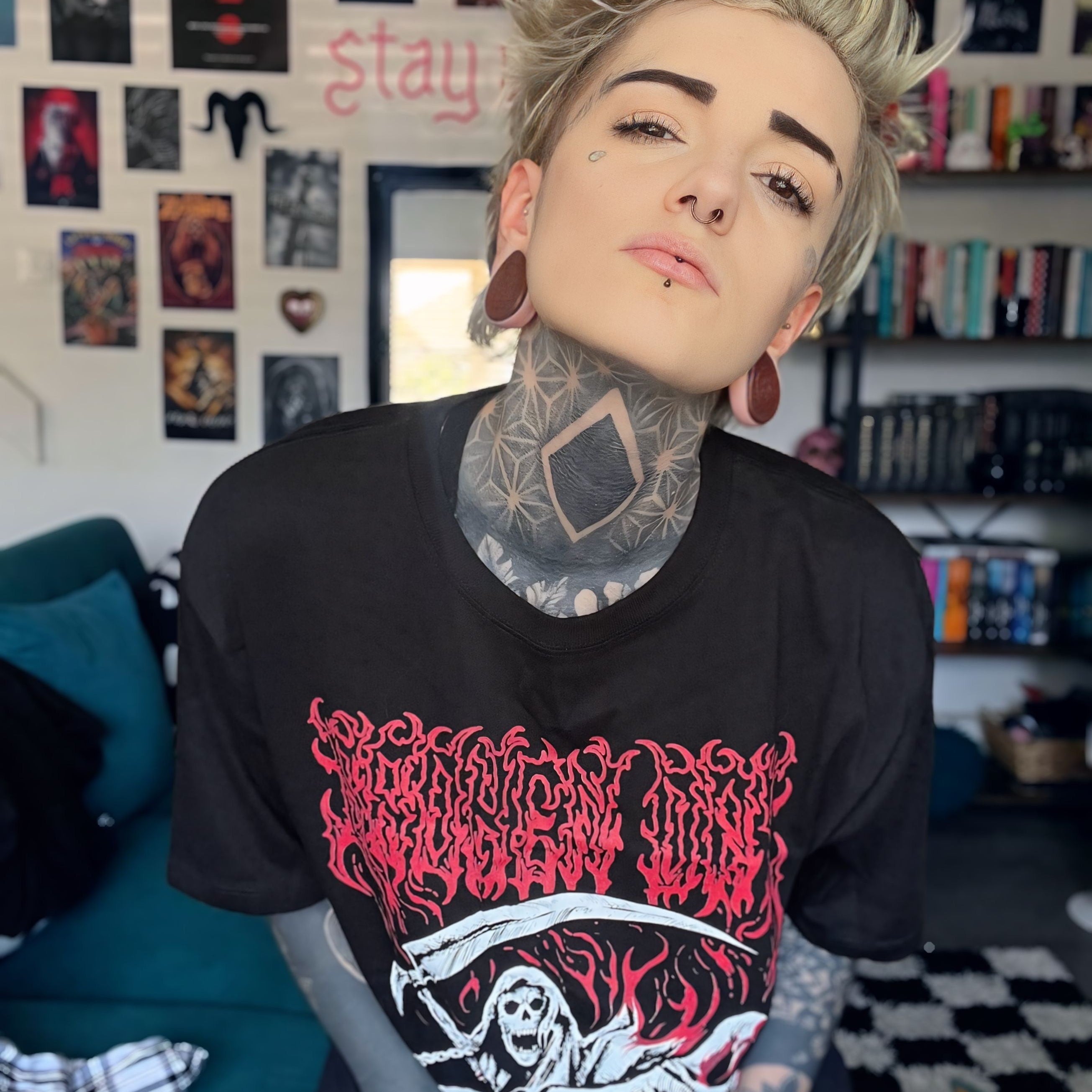 Person wearing a black t-shirt with red and white graphics in a room with posters and books on the wall.