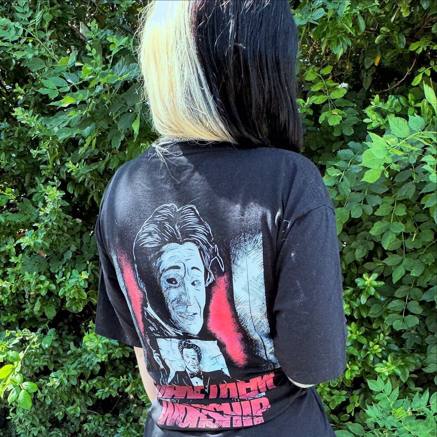 Person wearing a black t-shirt with graphic designs and text, standing in front of green foliage.