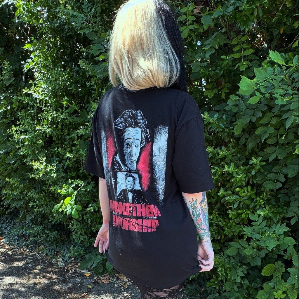 Person wearing a black t-shirt with graphic designs and text, standing outdoors with greenery in the background.