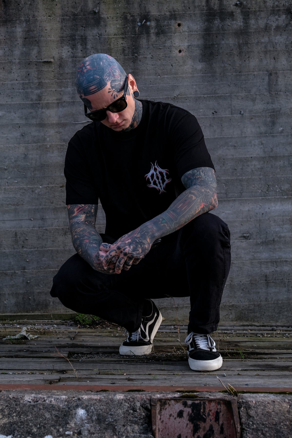 A tattooed man crouching in front of a concrete wall, wearing a black t-shirt with a white graphic logo on the chest, black pants, and black and white sneakers.